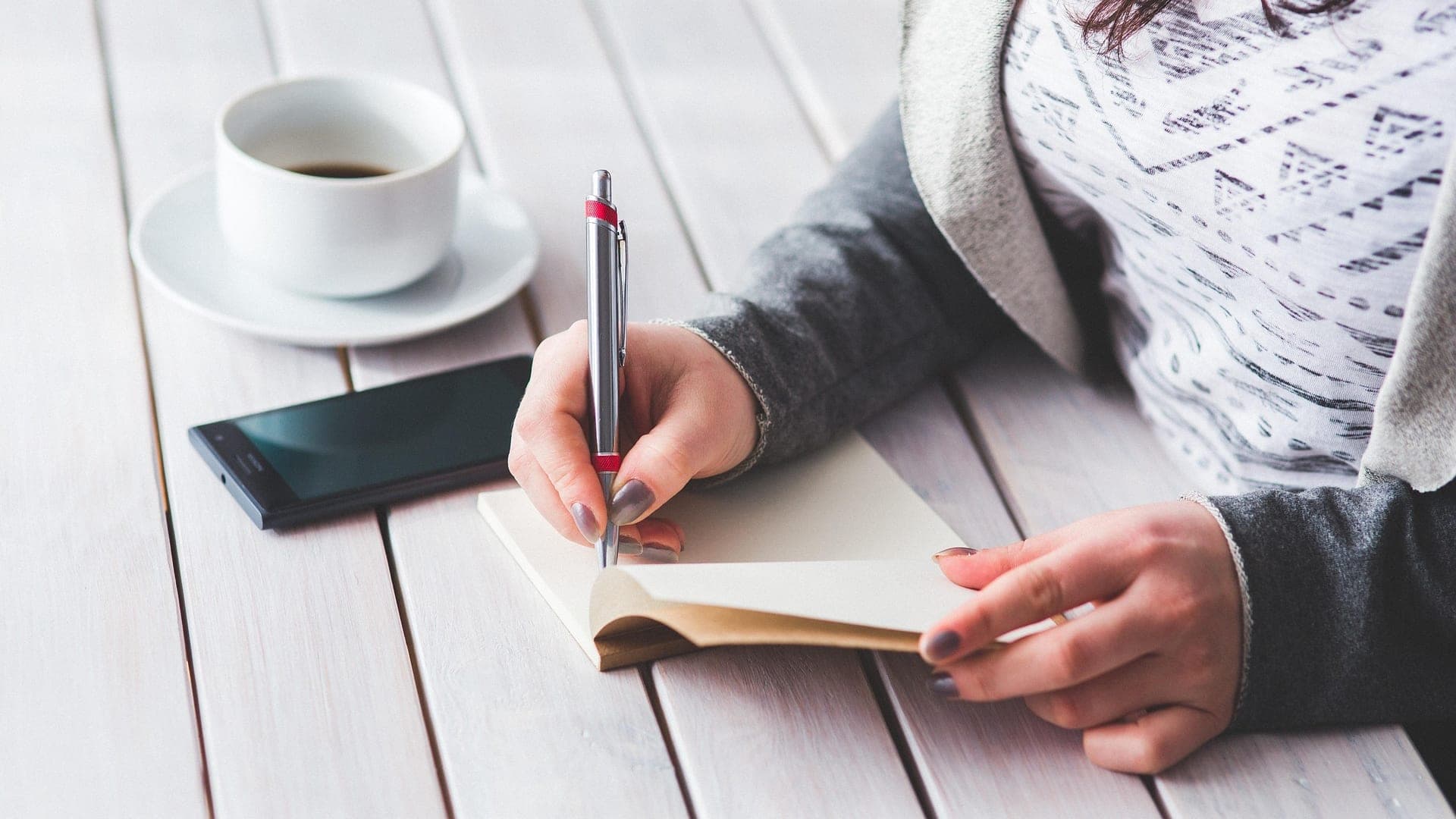 Mulher fazendo anotações num caderno, com café e celular sobre a mesa