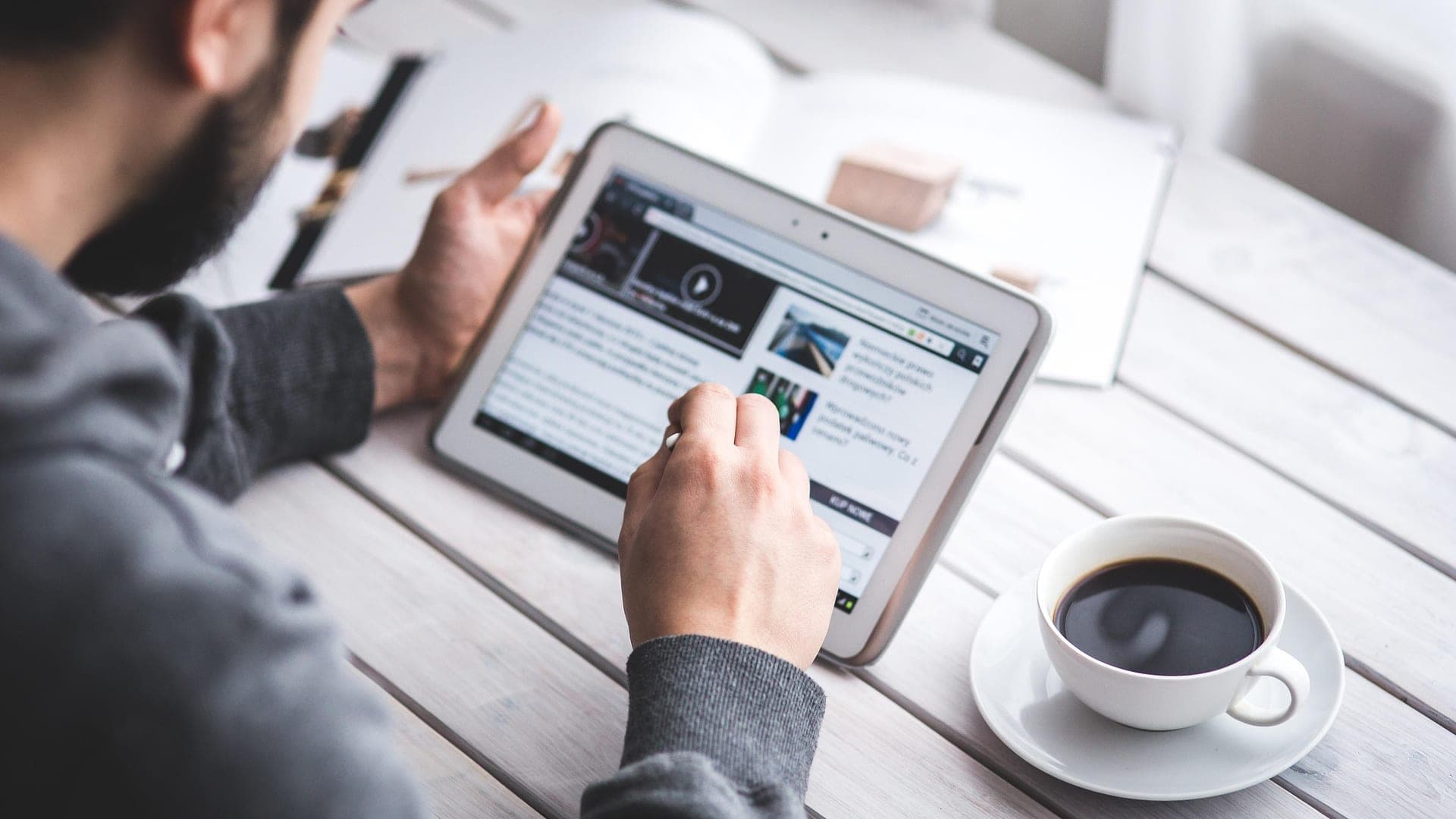 Homem usando um tablet numa mesa de madeira, ao lado de uma xícara de café
