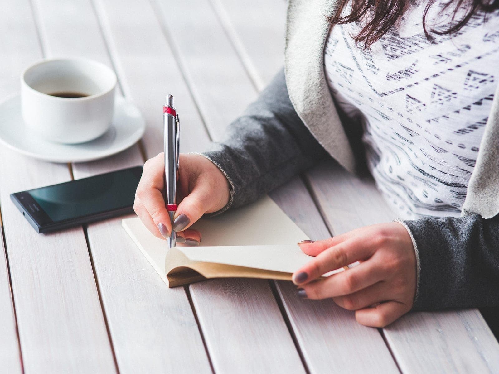 Mulher fazendo anotações num caderno, com café e celular sobre a mesa