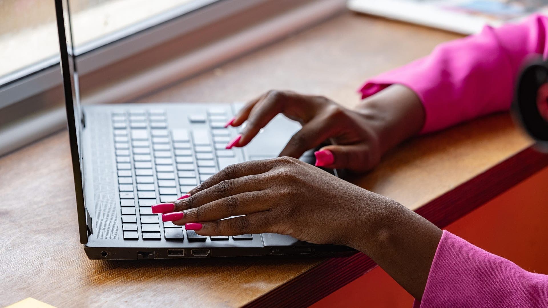 Mãos de uma mulher digitando no notebook, numa mesa perto da janela