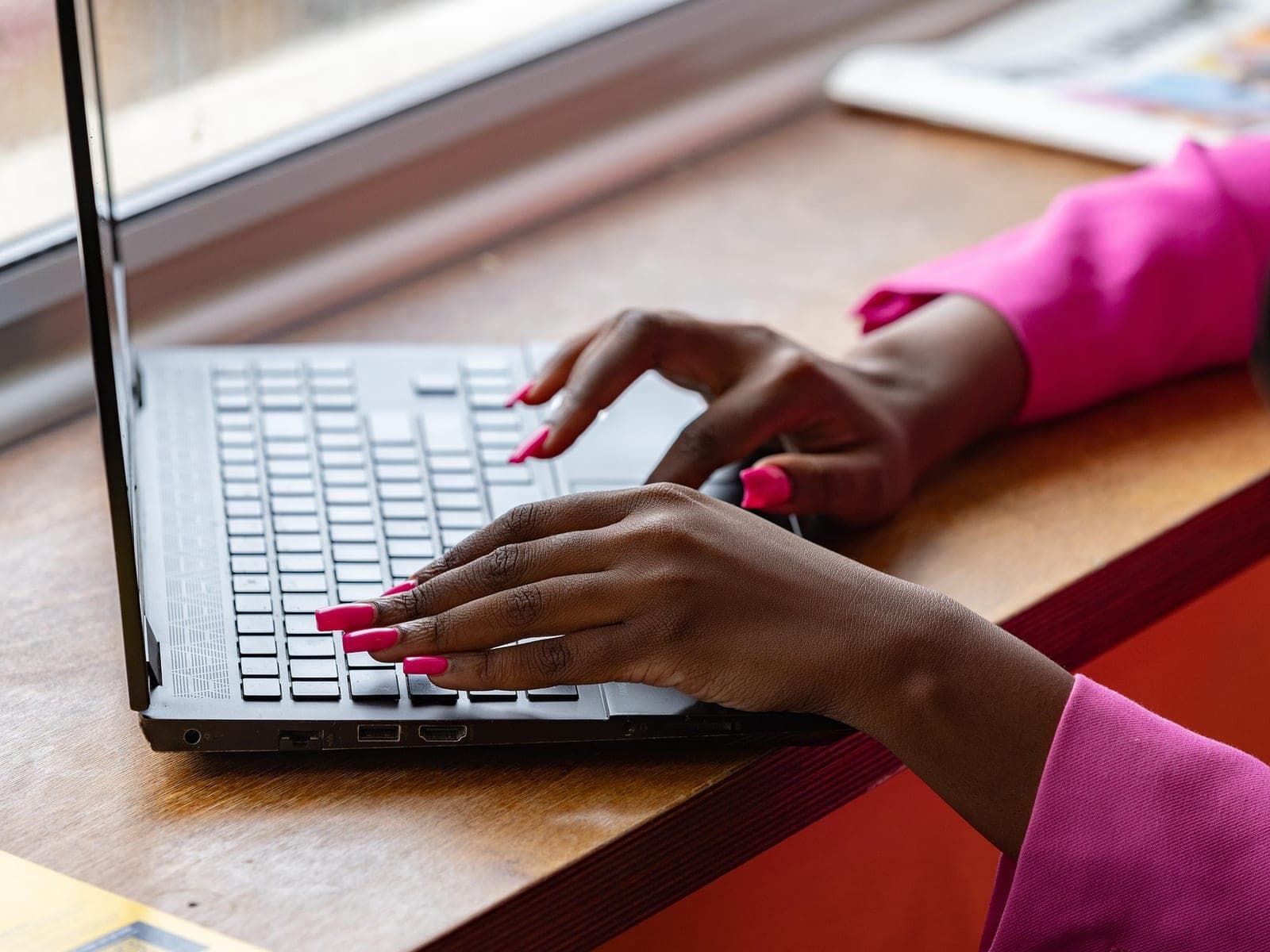 Mãos de uma mulher digitando no notebook, numa mesa perto da janela